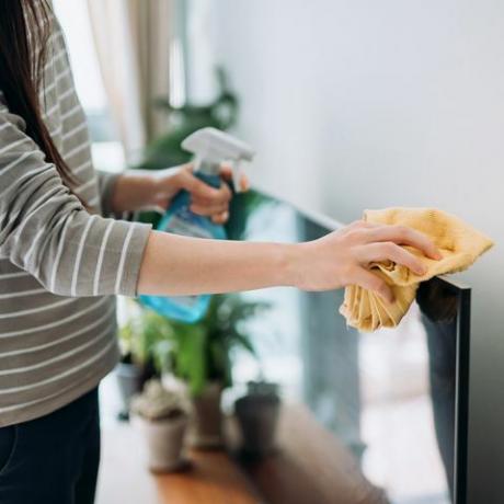 bidikan yang dipotong dari seorang wanita muda yang sedang membersihkan permukaan tv dengan semprotan pembersih dan kain antistatis di rumah pada siang hari