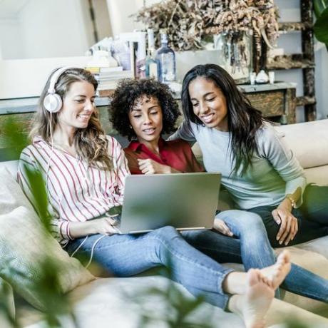 Tiga wanita bahagia dengan laptop duduk di sofa