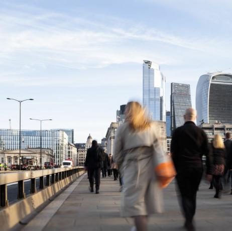 Inggris, london, tampilan belakang pria dan wanita bisnis yang berjalan ke tempat kerja melalui jembatan london dengan pemandangan distrik keuangan