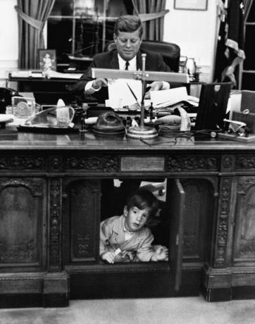 john f kennedy sedang bekerja di meja tegas di kantor oval Gedung Putih, sementara putranya, john f kennedy jr, bersembunyi di dalam perabotan