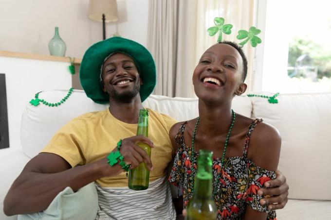 potret pasangan afrika amerika yang tertawa dengan kostum hari st patrick memegang botol bir tinggal di rumah dalam isolasi selama penguncian karantina