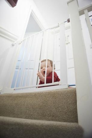 Gerbang pengaman bayi di tangga: Bayi laki-laki di pendaratan rumahnya. Dia berada di belakang gerbang keselamatan bayi yang menghentikannya dari bahaya tangga.